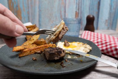 Elde çatal, hamburger bifteği, patates kızartması ve tabakta kızarmış yumurta.