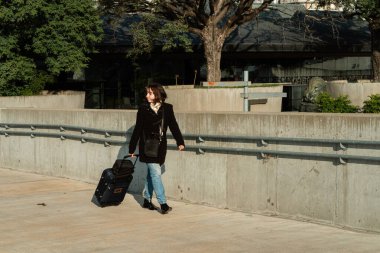 Güneşli bir günde geniş bir beton yolda yürüyen tekerlekli bavulu olan bir kadın. Kentsel hareket, seyahat ve bağımsızlık kavramı.