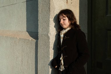 Gri bir duvara yaslanmış sert bir suratı olan genç bir kadın. Duygusal derinliği ve modern tarzı olan şehir portresi.