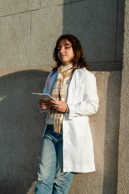 Genç bayan doktor tabletiyle kaldırımda yürüyor, hastane dışında günlük rutininin bir parçası. Etkin ve doğal görüntü.