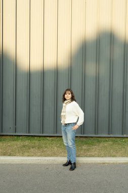 Genç bir kadın, büyük, koyu gri bir metal duvarın yanındaki parkta bir patikada duruyor. Görüntü minimalizmi ve insan ölçeğini çağrıştırıyor..