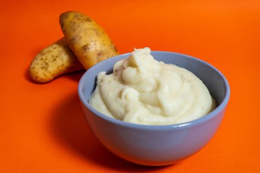 Yanında iki çiğ patatesle bir kase pürüzsüz patates püresi canlı turuncu bir zemin karşısında görsel olarak çekici bir tezat oluşturuyor.