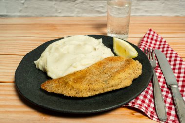 Patates püresi ve limon dilimiyle servis edilen balıklı fileto, ahşap bir masada siyah bir tabakta sunuluyor.