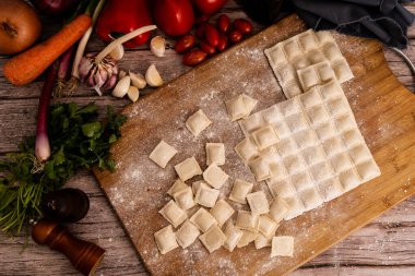 Ahşap bir tahtaya un serpilmiş, sarımsak, domates ve maydanozla pişirilmeye hazır, pişirilmemiş ravioli makarna parçaları.