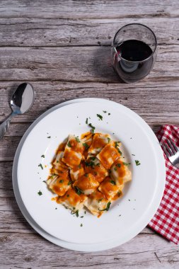 Lezzetli domates soslu ravioli makarna, taze maydanozla serpilmiş, beyaz bir tabakta servis ediliyor, yanında da ahşap bir masada kırmızı şarap var.