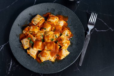 Domates soslu ve maydanozlu lezzetli ravioli makarnası, siyah tabakta servis edilir, görsel olarak çekici bir mutfak sahnesi yaratır.