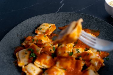 Lezzetli ravioli makarnası, canlı domates sosuyla servis edilir ve modern koyu gri tabakta taze maydanozla süslenir.