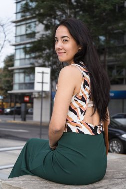 Uzun siyah saçlı, genç bir Latin kadın bir şehir sokağında bankta oturuyor ve arkasını kolluyor.