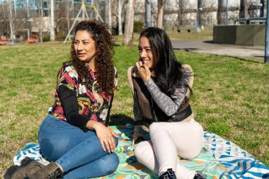 İki bayan arkadaş güneşli bir günün tadını çıkarıyor, bir şehir parkında battaniyede oturuyor, konuşuyor ve birlikte gülüyorlar.