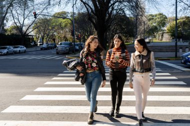Üç kız öğrenci yaya geçidinde karşıdan karşıya geçerken yürüyüp sohbet ediyorlar.