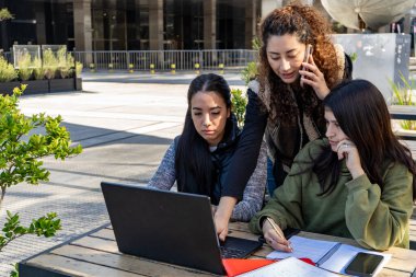 Üç üniversite öğrencisi açık havada bir projede iş birliği yapıyor, bir dizüstü bilgisayar kullanıyor ve bir öğrenci telefonla konuşurken not alıyor.