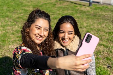 Parkta akıllı bir telefonla selfie çeken iki mutlu bayan arkadaş, açık havada güneşli bir günün tadını çıkarıyorlar.
