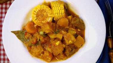 Şef, kırmızı kareli masa örtüsüne mısır, et ve sebze içeren doyurucu bir tencere güveç koyar.