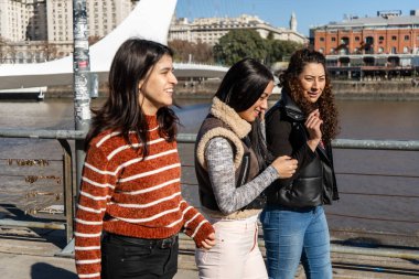 Güneşli bir günün tadını çıkaran üç genç kadın, Buenos Aires 'in Puerto Madero bölgesinde nehri kenarında sohbet edip gülüyorlar.