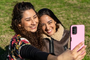 Parkta akıllı bir telefonla selfie çeken iki mutlu bayan arkadaş, açık havada güneşli bir günün tadını çıkarıyorlar.