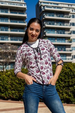 Modern apartmanların önünde elleri ceplerinde duran kendine güvenen genç bir kadın, şehir yaşamının tadını çıkarıyor.