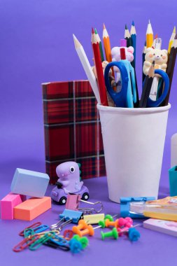 Renkli kalemler, makas, silgi ve klipler gibi kırtasiye malzemeleri mor bir arkaplan üzerinde düzenlenmiş, canlı bir okula dönüş sahnesi oluşturur