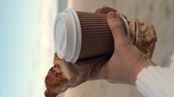 品尝新鲜的羊角面包 咖啡在纸杯女性手中的海滩海背景 户外早餐福利的概念 Fpv垂直 带走食物 咖啡休息的替代办法 — 图库视频影像