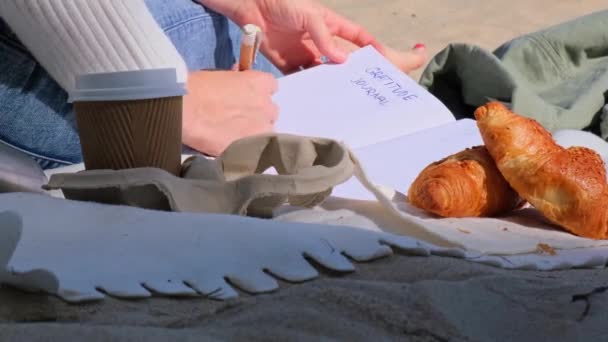 海の景色の隣のビーチでノートを持つ若い学生の研究 コーヒーを飲み クロワッサンを食べる 感謝日記自己反省自己発見を書く屋外暖かい秋の海岸 — ストック動画