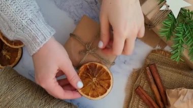 Yeni yıl hediyeleriyle dolu bir kutu hazırlayan kadın. Kağıda sarılmış ve kuru portakal dilimleriyle süslenmiş. Tatiller ve Hediyeler konsepti. El yapımı çevre dostu alternatif yeşil Noel hediyeleri sıfır atık