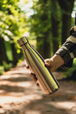 Tanımlanamayan bir kadının su şişesini tuttuğu yakın plan. Parkta yeniden kullanılabilir çelik termo su şişesi. Sürdürülebilir yaşam tarzı. Plastiksiz sıfır atıksız yaşam. Yeşil Çevre Koruma 'ya git