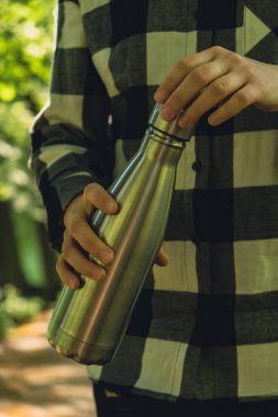 Tanımlanamayan bir kadının su şişesini tuttuğu yakın plan. Parkta yeniden kullanılabilir çelik termo su şişesi. Sürdürülebilir yaşam tarzı. Plastiksiz sıfır atıksız yaşam. Yeşil Çevre Koruma 'ya git