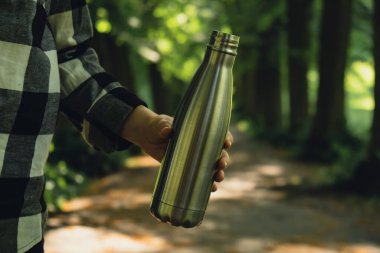 Tanımlanamayan bir kadının su şişesini tuttuğu yakın plan. Parkta yeniden kullanılabilir çelik termo su şişesi. Sürdürülebilir yaşam tarzı. Plastiksiz sıfır atıksız yaşam. Yeşil Çevre Koruma 'ya git