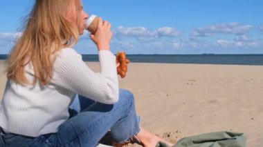 Kâğıt bardaktan kahve içen mutlu genç bir kadın sahildeki okyanusta kruvasan yiyor. Sonbahar bulutlu gününün tadını çıkar. Doğada rahatlık. Sakinlik ve rahatlama konsepti. Seyahat kavramı