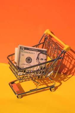 Amerikan doları kağıt para birimi 5 banknot para renkli turuncu arka planda alışveriş arabası. Metnin için alanı kopyala. Çevrimiçi alışveriş, alışveriş merkezi market tüketicisi konsepti. Küçük oyuncak