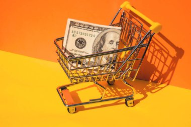 Amerikan doları kağıt para birimi 100 banknot para renkli turuncu arka planda alışveriş arabası. Metnin için alanı kopyala. Çevrimiçi alışveriş, alışveriş merkezi market tüketicisi konsepti. Küçük oyuncak