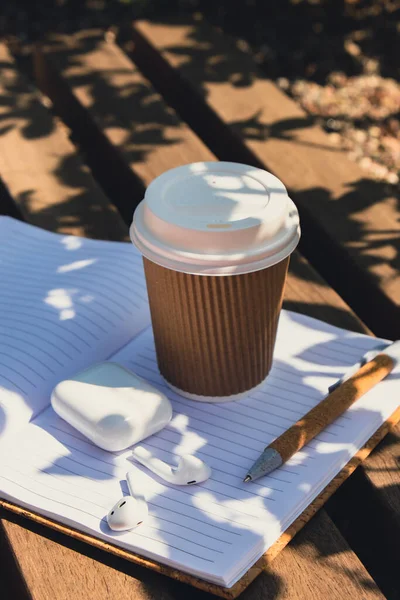 Take away coffee in craft recycling paper cup with paper notebook with wireless headphones. Mockup Coffee break. Audio healing, sound therapy wellness rituals, positive mental health habits listening