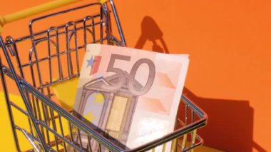 Sarı arka planda oyuncak süpermarket tramvayına 50 Euro 'luk banknot ekle. Satış sepeti dolusu euro. Asgari yaşam maaşı kavramı: kredi, yatırım emekliliği tasarruf finansmanı
