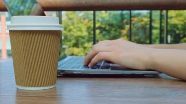 Tanınmayan genç bir kadın alışveriş merkezindeki ahşap masada modern bir dizüstü bilgisayarda çalışıyor. Kağıt bardaktan kahve içmek. Eş zamanlı çalışma alanında klavyede yazan Öğrenci Çalışan Kadın Elleri