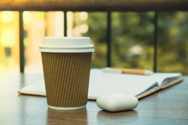 Hot latte coffee in craft recycling paper cup with paper notebook with wireless headphones. A take away paper cup on cafe table. Freelance Workspace notebook with coffee for productive work and study