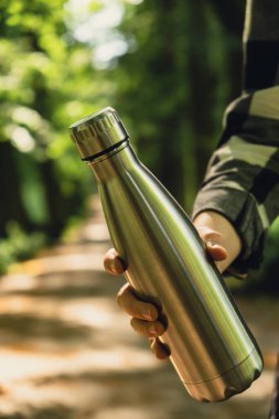 Tanımlanamayan bir kadının su şişesini tuttuğu yakın plan. Parkta yeniden kullanılabilir çelik termo su şişesi. Sürdürülebilir yaşam tarzı. Plastiksiz sıfır atıksız yaşam. Yeşil Çevre Koruma 'ya git