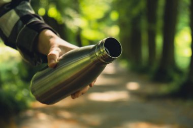 Tanımlanamayan bir kadının su şişesini tuttuğu yakın plan. Parkta yeniden kullanılabilir çelik termo su şişesi. Sürdürülebilir yaşam tarzı. Plastiksiz sıfır atıksız yaşam. Yeşil Çevre Koruma 'ya git