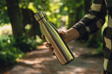 Tanımlanamayan bir kadının su şişesini tuttuğu yakın plan. Parkta yeniden kullanılabilir çelik termo su şişesi. Sürdürülebilir yaşam tarzı. Plastiksiz sıfır atıksız yaşam. Yeşil Çevre Koruma 'ya git