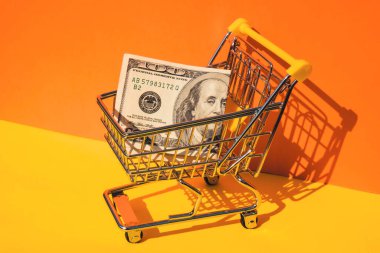 Amerikan doları kağıt para birimi 100 banknot para renkli turuncu arka planda alışveriş arabası. Metnin için alanı kopyala. Çevrimiçi alışveriş, alışveriş merkezi market tüketicisi konsepti. Küçük oyuncak