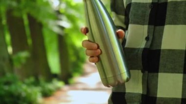 Tanımlanamayan bir kadının su şişesini tuttuğu yakın plan. Parkta yeniden kullanılabilir çelik termo su şişesi. Sürdürülebilir yaşam tarzı. Plastiksiz sıfır atıksız yaşam. Yeşil Çevre Koruma 'ya git