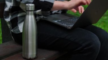 Su şişesi. Ahşap bankta yeniden kullanılabilir çelik termo su şişesi. Sürdürülebilir yaşam tarzı. Plastiksiz sıfır atıksız yaşam. Yeşil çevre korumasına geçin. Dizüstü bilgisayarla çalışan bir kadın öğrenci.