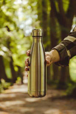 Tanımlanamayan bir kadının su şişesini tuttuğu yakın plan. Parkta yeniden kullanılabilir çelik termo su şişesi. Sürdürülebilir yaşam tarzı. Plastiksiz sıfır atıksız yaşam. Yeşil Çevre Koruma 'ya git