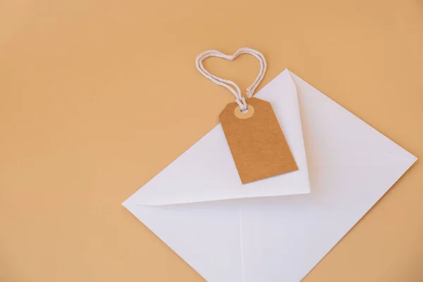 White envelope with beige paper note and rope in heart shape on neutral beige background. Greeting card holiday idea. Mock up copy space for text. Gift present