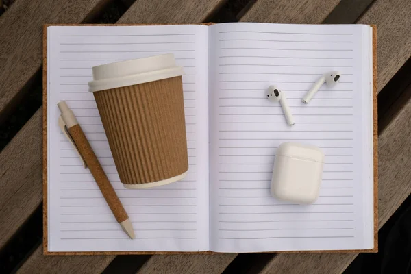 Take away coffee in craft recycling paper cup with paper notebook with wireless headphones. Mockup Coffee break. Audio healing, sound therapy wellness rituals, positive mental health habits listening podcast writing self discovery gratitude journal.