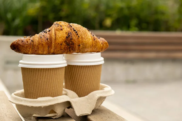 Fresh baked chocolate croissant on Two paper cups with lid for tea to go. Breakfast Coffee take away on the table. Take-out coffees with brown paper cup holder. Brown safety cardboard collars. Take