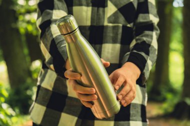 Close-up of Unrecognizable female hand holding Water bottle. Reusable steel thermo water bottle in park. Sustainable lifestyle. Plastic free zero waste free living. Go green Environment protection. Woman drinking water in park 