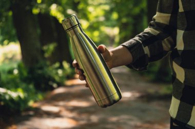 Close-up of Unrecognizable female hand holding Water bottle. Reusable steel thermo water bottle in park. Sustainable lifestyle. Plastic free zero waste free living. Go green Environment protection. Woman drinking water in park 
