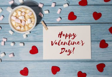 HAPPY VALENTINES DAY text Greeting card with red envelope with white cup of coffee and marshmallows on wooden background. Romantic Small hearts Valentine day. Holiday morning. Top view, flat lay minimalist