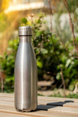 Water bottle. Reusable steel thermo water bottle on wooden bench. Sustainable lifestyle. Plastic free zero waste free living. Go green Environment protection. Health-conscious. Steel thermo water bottle of silver, on background of blurred grass. 