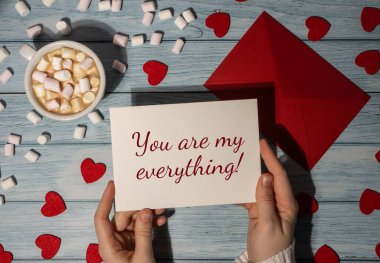 YOU ARE MY EVERYTHING valentine text Female hands holding valentine card red envelope with white cup of coffee and marshmallows on wooden blue background. Romantic Small hearts Valentine day. Holiday morning. Top view, flat lay minimalist branding co