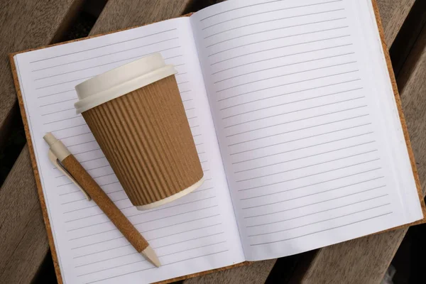 Eco recycling paper cup with coffee or tea on kraft paper with empty paper notebook on wooden bench. Concept of study work outdoors. Take away coffee to go. Copy space for text. Disposable Cardboard coffee outdoors